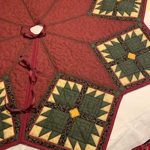 May include: A quilted Christmas tree skirt with a burgundy background and green and gold star-shaped blocks. The skirt has a red ribbon tied in a bow at the center.