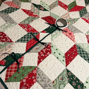 May include: A quilted Christmas tree skirt with a starburst design. The skirt features red, green, and white patterned fabric pieces, including snowflakes and plaid. A dark green ribbon is attached for securing the skirt around a tree.