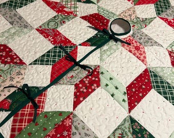 May include: A quilted Christmas tree skirt with a starburst design. The skirt features red, green, and white patterned fabric pieces, including snowflakes and plaid. A dark green ribbon is attached for securing the skirt around a tree.