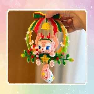 May include: A pink and white beaded Christmas ornament shaped like a cartoon animal wearing a Santa hat and holding a candy cane. The ornament is hanging from a green and yellow beaded wreath with a red bow and a yellow bell.