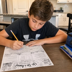 May include: A person is seated at a wooden table, writing on a worksheet with a pencil. The worksheet has the text "Up, Up & Away!" and illustrations. A blue lunch bag and notebooks are on the table.