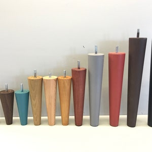 Op de afbeelding: Assortiment van 12 houten meubellepoten in verschillende kleuren en afwerkingen, waaronder naturel hout, bruin, grijs, rood en teal. De poten zijn taps toelopend en hebben een schroefdesign.