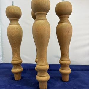 May include: Four unfinished wooden furniture legs. Each leg has a turned design with a bulbous top and bottom, and a threaded metal rod extending from the top. The legs are a light brown colour and are displayed against a blue background.