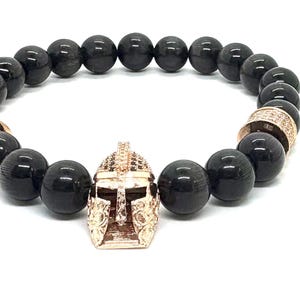 May include: Black beaded bracelet with gold-colored accents. The bracelet features large, round, black beads, two gold-colored spacers, and a gold-colored Spartan helmet charm. The bracelet is displayed on a white background.