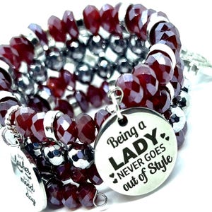 May include: A multi-strand bracelet featuring faceted burgundy beads, silver beads, and silver accents. The bracelet includes round silver charms with the phrases "Good lashes, good mood, good day" and "Being a LADY NEVER GOES out of Style."