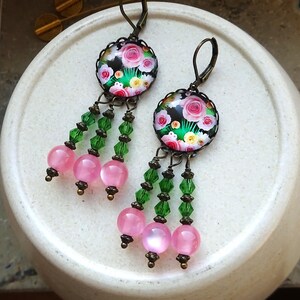 Op de afbeelding: Een paar hangende oorbellen met ronde cabochons met een bloemenpatroon van roze en gele rozen op een zwarte achtergrond. Elke oorbel heeft drie hangende strengen van groene gefacetteerde kristalkralen die eindigen in gladde, ronde roze kralen.