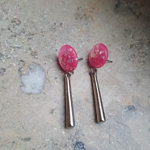 May include: A pair of dangle earrings with pink and silver accents. The earrings feature a pink oval-shaped top with silver glitter and a long, thin silver dangle.