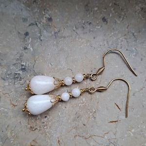 May include: White teardrop-shaped earrings with gold accents and white beads. The earrings are made of a white stone and have a gold hook.