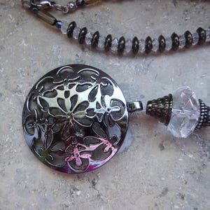 Könnte beinhalten: Ein silberfarbener Anhänger mit floralem Filigranmuster, Teil einer Halskette. Die Halskette hat klare und dunkle Perlen, mit einem dekorativen Kristallelement. Der Schmuck ist vor einem neutralen Hintergrund platziert.