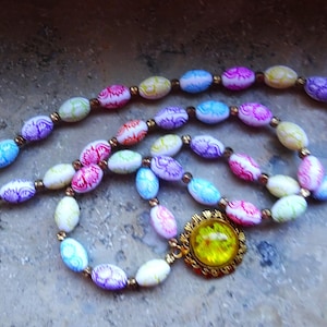 May include: A colorful necklace featuring oval-shaped beads in shades of pink, purple, blue, and green. The beads have a floral pattern. The necklace has a gold-colored clasp and a gold-framed pendant with a yellow center.