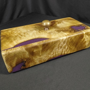 May include: A rectangular wooden box featuring a polished light brown burl wood grain with natural edges filled with opaque purple resin. The lid includes a centered, small, round, metallic gold-toned knob handle for opening the container.