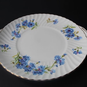 Peut inclure: Assiette de service en céramique blanche avec un bord festonné et une bordure dorée. L'assiette est ornée de motifs floraux bleus. L'assiette a deux poignées et est posée sur un fond sombre.