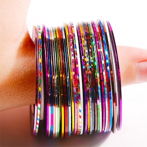Peut inclure: Un rouleau de ruban adhésif pour nail art de différentes couleurs. Le ruban est fin et métallique, avec une variété de couleurs et de finitions, y compris l'or, l'argent, le rouge, le bleu, le vert et le violet.