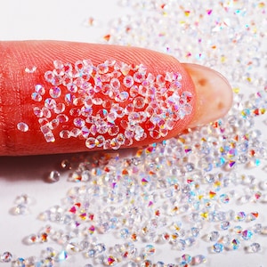 Peut inclure: Gros plan sur un bout de doigt recouvert de strass clairs irisés. Les strass sont éparpillés sur une surface blanche.
