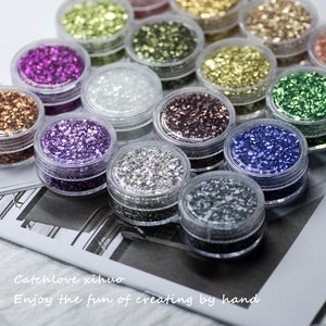 Puede incluir: Surtido de pequeños frascos llenos de purpurina de varios colores. Los frascos están dispuestos en filas sobre una superficie blanca. El texto "Catchlove xihua Enjoy the fun of creating by hand" es visible en la parte inferior de la imagen.