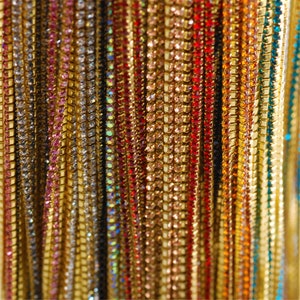 May include: A close-up of a variety of colourful rhinestone chains. The chains are made of small, sparkling rhinestones that are strung together on a thin, gold-coloured wire. The chains are arranged in a vertical pattern, creating a shimmering and eye-catching display.
