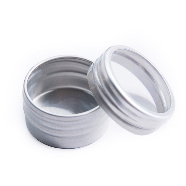 May include: A silver metal container with a clear lid. The container is round and has a smooth surface. The lid has a slightly raised edge.