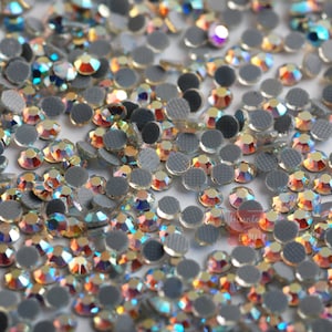 May include: A close-up view of a large quantity of iridescent, round, flat-backed rhinestones. The rhinestones have a silver backing and are scattered on a white surface.