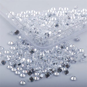 May include: A close-up of a clear plastic container filled with loose, clear, round rhinestones. The container is sitting on a white surface and some rhinestones have spilled out.