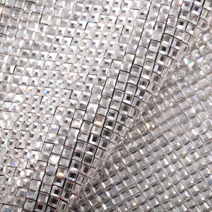 May include: Close-up of a silver sparkly fabric with a diamond-like pattern. The fabric is made of small, square-shaped sequins that are sewn together.