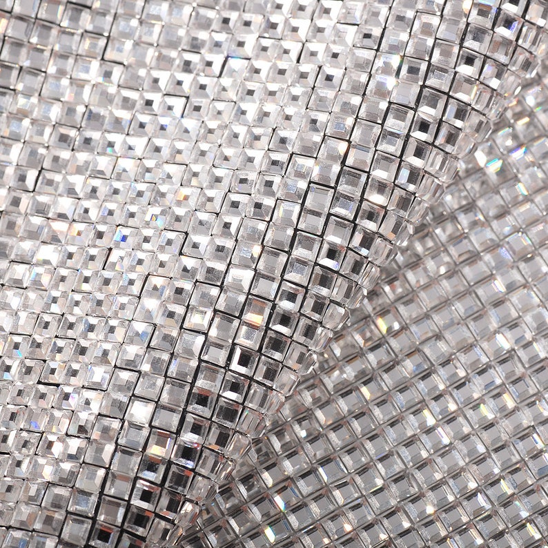 May include: Close-up of a silver sparkly fabric with a diamond-like pattern. The fabric is made of small, square-shaped sequins that are sewn together.