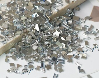 May include: A collection of silver-colored, reflective rhinestones in various shapes, including butterflies, hearts, and geometric forms. The rhinestones are scattered on a white surface, with a piece of beige material in the background. The image is well-lit, highlighting the sparkle of the rhinestones.