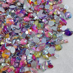 May include: Assortment of iridescent rhinestones in various shapes and sizes, including round, oval, and square. The rhinestones are in shades of pink, purple, and blue, with a rainbow-like shimmer.