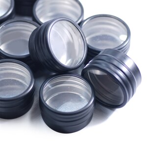 May include: A group of black metal containers with clear lids. The containers are round and have a ribbed design. They are stacked on top of each other.