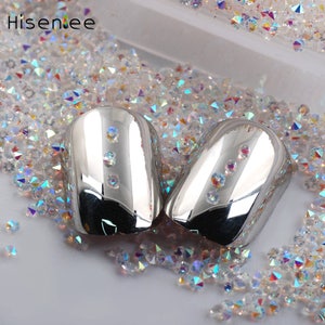 Puede incluir: Dos puntas de uñas de color plateado con un acabado de alto brillo, rodeadas de diamantes de imitación iridiscentes.