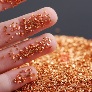 Può includere: Primo piano di una mano che tiene piccoli pezzi irregolari di foglia d'oro. La foglia d'oro è sparsa su una superficie, creando un effetto scintillante e metallico.