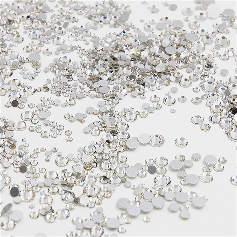 May include: A large assortment of clear glass rhinestones of various sizes. The rhinestones are scattered on a white surface.