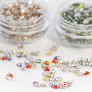 Puede incluir: Surtido de piedras de cristal facetadas en forma de corazón, de colores, esparcidas sobre una superficie blanca. Las piedras de cristal son de varios tonos de rojo, naranja, amarillo, verde, azul y morado.