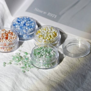 Pode incluir: Quatro pequenos frascos transparentes cheios de contas iridescentes coloridas. Os frascos estão sobre uma superfície de tecido branco. As contas são azuis, laranja, amarelas e verdes.