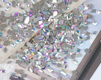 May include: A collection of iridescent rhinestones in various shapes, including diamonds, ovals, and stars. The rhinestones have a silver backing and reflect multiple colors. They are scattered on a light-colored surface.