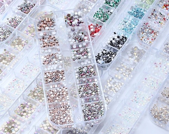 Pode incluir: Uma coleção de recipientes de plástico transparente cheios de strass de várias cores. Os strass são em tons de rosa, transparente, castanho, verde e preto. Os recipientes estão dispostos em um padrão de grade, ideal para artesanato e nail art.