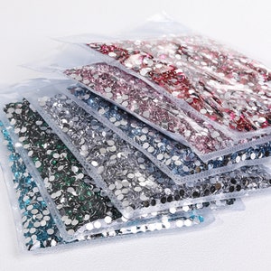 K&ouml;nnte beinhalten: Sortiment aus durchsichtigen Plastikt&uuml;ten gef&uuml;llt mit verschiedenen Farben runder Strasssteine. Die Farben umfassen blaugr&uuml;n, silber, hellblau, dunkelblau, rosa und rot.