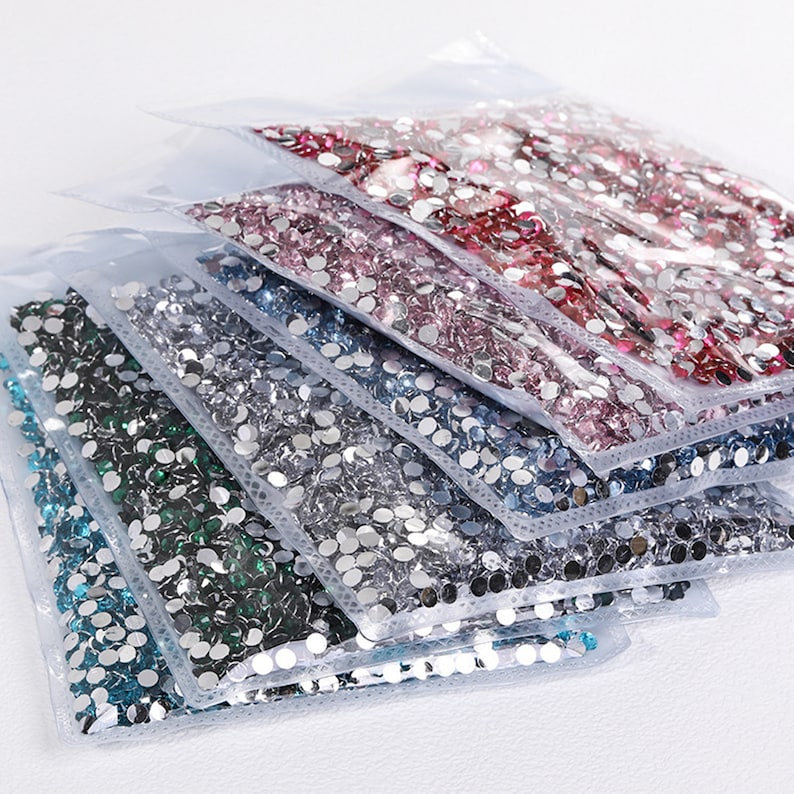 K&ouml;nnte beinhalten: Sortiment aus durchsichtigen Plastikt&uuml;ten gef&uuml;llt mit verschiedenen Farben runder Strasssteine. Die Farben umfassen blaugr&uuml;n, silber, hellblau, dunkelblau, rosa und rot.