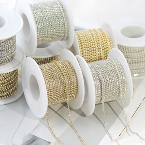 May include: Spools of rhinestone trim in various colors, including silver, gold, and iridescent. The trim is made of small, sparkling rhinestones set in a chain. The spools are white and arranged on a white surface.