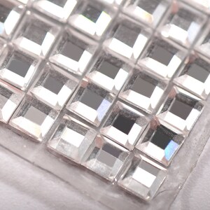 May include: Close-up of a sheet of clear, square-shaped rhinestones. The rhinestones are arranged in a grid pattern and have a reflective surface.