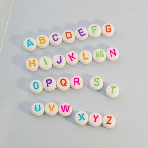 以下が含まれることがあります： カラフルな文字が書かれた白いアルファベットのビーズ。ビーズはAからZまでのアルファベット順に並んでいます。