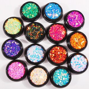 Può includere: Dodici piccoli contenitori rotondi pieni di glitter colorati e iridescenti. Ogni contenitore ha un coperchio trasparente ed è pieno di un colore di glitter diverso.