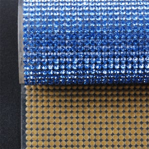 Può includere: Un rotolo di passamaneria di strass blu con un retro adesivo trasparente. La passamaneria è su un foglio di supporto bianco. La passamaneria è mostrata accanto a un rotolo di passamaneria di strass dorata con un retro adesivo trasparente.