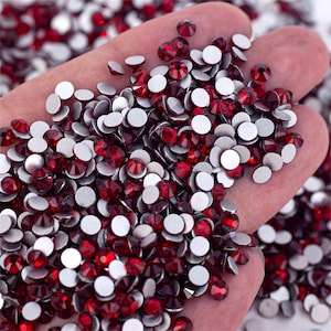 May include: A close-up of a hand holding a large quantity of red rhinestones with silver backs. The rhinestones are scattered and overlapping.