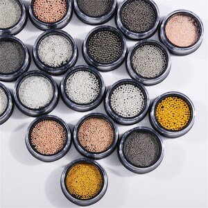 May include: Assortment of small, round, metallic beads in various colors, including silver, gold, and rose gold, displayed in small, clear containers with black lids.