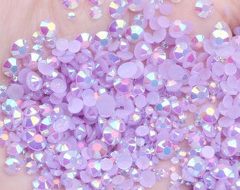 May include: A close-up of a large assortment of iridescent lavender rhinestones of various sizes. The rhinestones are scattered on a hand, creating a sparkling and glamorous effect.