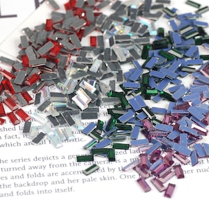 Puede incluir: Una colección de brillantes rectangulares iridiscentes en varios colores, incluyendo rojo, plateado, transparente, verde, azul y morado. Estas pequeñas gemas decorativas están dispersas sobre una superficie blanca, ideales para manualidades.