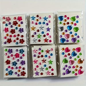 May include: Six packs of colorful, self-adhesive rhinestone stickers. Each pack contains a variety of shapes, including flowers, stars, and hearts, in shades of red, pink, blue, green, purple, and gold. The stickers are arranged on white backing cards.