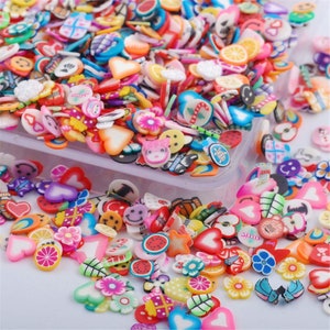May include: A variety of colorful, small, plastic charms in the shape of hearts, flowers, fruits, and other designs. The charms are scattered on a white surface.