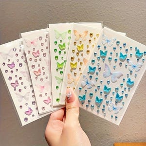 May include: A collection of butterfly stickers in various colors, including purple, pink, green, yellow, and blue. Each sheet features butterfly designs and small, sparkling accents. The stickers are displayed on a white background.