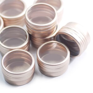 May include: A group of small, round, gold-coloured metal containers with clear lids. The containers are stacked and scattered on a white surface.